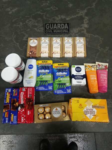 Imagem mostrando produtos diversos, incluindo chocolates, itens de higiene pessoal e cremes, dispostos sobre uma superfície. No fundo, há uma placa escrita 'GUARDA CIVIL MUNICIPAL'.