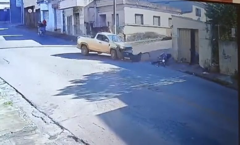 Uma cena de rua mostrando um carro amarelo estacionado em uma calçada, com uma pessoa próxima a ele e algumas casas ao fundo.