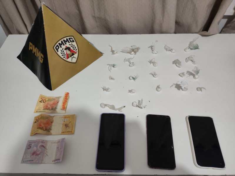 Pyramid com o logotipo da PMMG, três smartphones pretos e um branco, dinheiro em notas de R$ 5 e R$ 20, e pequenos papéis em uma mesa.