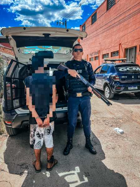 Policial de uniforme segurando uma arma ao lado de um suspeito algemado em uma área urbana.