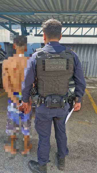 Um policial da Guarda Civil Municipal em pé, observando um homem algemado e descalço, em uma área externa.