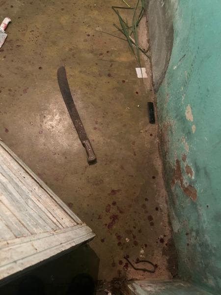 Um ambiente interior com um chão sujo e manchas de sangue. Um facão está deitado no chão, ao lado de um objeto escuro e uma folha de grama.