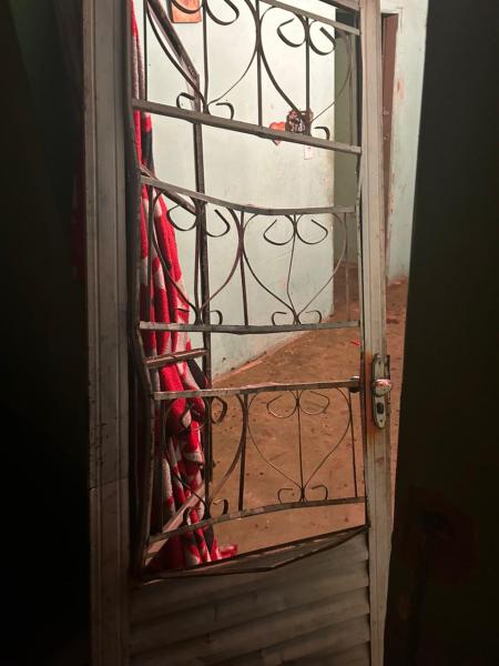 Uma porta de metal com grade aberta, mostrando um interior simples, com chão de terra e paredes sem revestimento. Atrás da porta, há um pano vermelho pendurado.