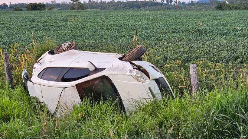 Um carro branco capotado em um campo verde, com a roda para cima e cercado por grama alta.