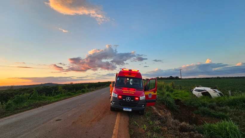 Ambulância da corporação de resgate parada ao lado da estrada, com as luzes de emergência ligadas, enquanto um carro acidentado está fora da pista ao fundo, sob um céu colorido de pôr do sol.