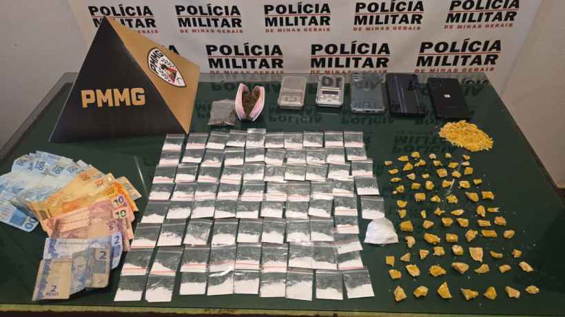 Imagem mostrando uma mesa com dinheiro em notas e diversas sacolas de drogas, além de celulares e equipamentos associados, com fundo de uma parede da polícia militar.