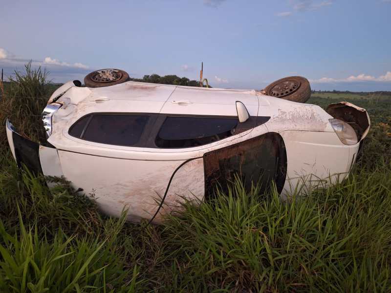 Carro branco capotado em meio à grama alta.