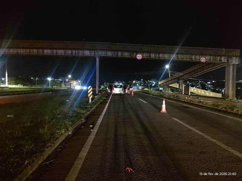 Foto de uma rodovia à noite, com sinalização de trânsito e cones de alerta, abaixo de uma ponte.