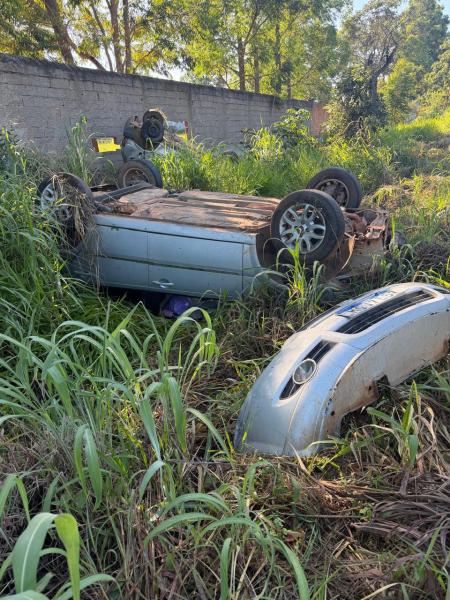 Carro deitado em um terreno coberto de grama alta, com um para-choque próximo e outros carros visíveis ao fundo.