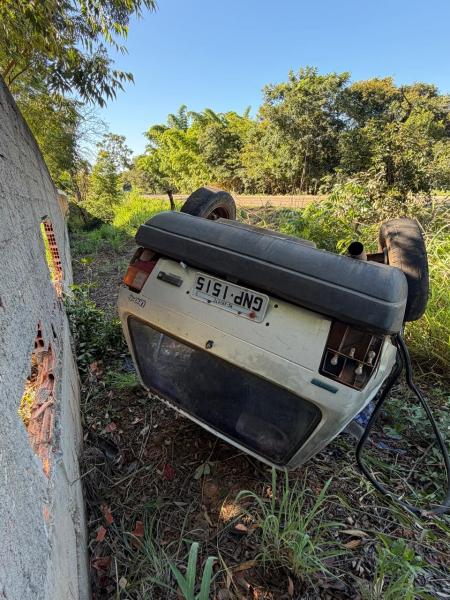 Um carro capotado próximo a uma parede de concreto, cercado por vegetação e árvores ao fundo.