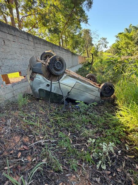 Veículo capotado em uma área verde ao lado de uma parede de concreto.