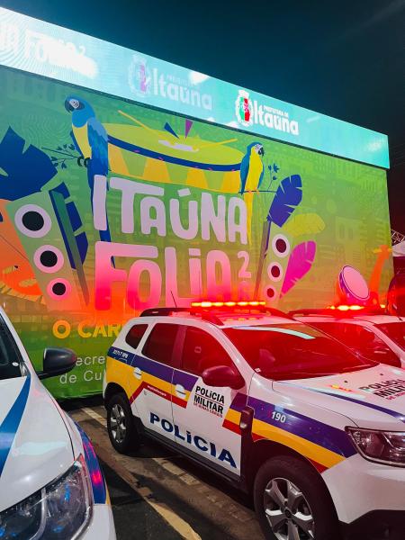 Veículo da Polícia Militar em primeiro plano, com um grande painel colorido ao fundo promovendo o evento "Itaúna Folia 2".