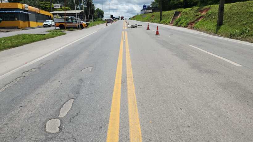 Vista de uma estrada com marcas de faixas amarelas, cones de sinalização e buracos na pista, com vegetação à direita e estabelecimentos comerciais à esquerda.