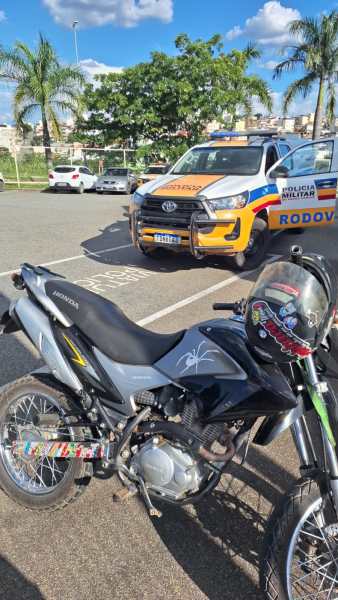 Uma motocicleta preta estacionada em frente a um veículo da polícia militar rodoviária em uma área pública com árvores ao fundo.
