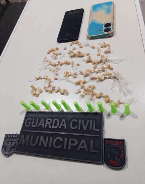 Imagem mostrando dois celulares, um preto e outro azul com detalhes amarelados, sobre uma mesa. Há uma quantidade de pequenos fragmentos em forma de arroz e vários objetos plásticos verdes. Também está visível uma placa que diz 'GUARDA CIVIL MUNICIPAL'.