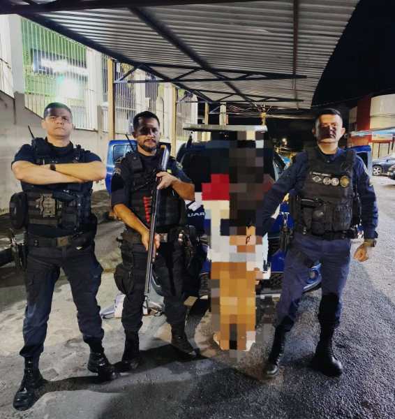 Três policiais em trajes táticos posando em uma rua, com um veículo de polícia ao fundo e uma pessoa detida ao centro, com partes do corpo desfocadas.