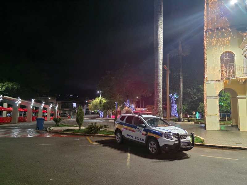 Veículo da polícia estacionado em uma praça iluminada à noite, com árvores decoradas e tendas vermelhas ao fundo.