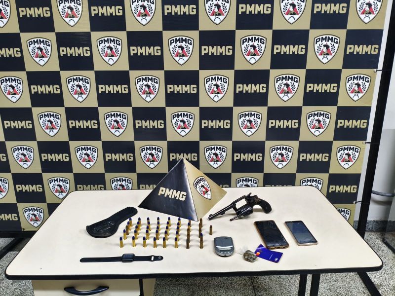 Mesa com uma arma de fogo, cartuchos, três celulares, um relógio e uma base decorativa com o logo da PMMG, em frente a um painel com várias imagens da PMMG.