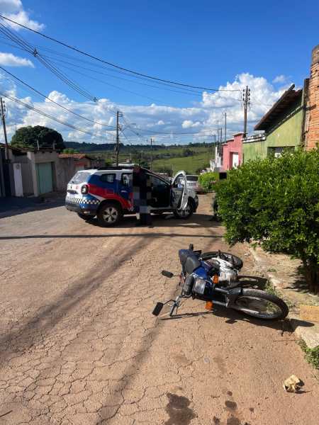 Uma moto caída ao lado de um carro de polícia com a porta aberta em uma rua tranquila. Ao fundo, um céu azul com nuvens e casas ao longo da rua.