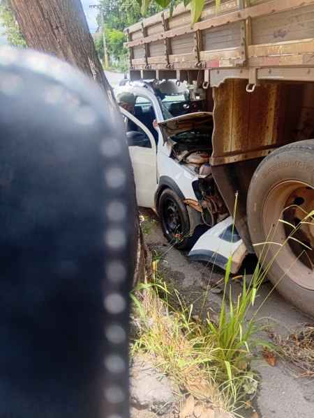 Acidente de carro envolvendo um veículo branco colidido embaixo de um caminhão, com danos visíveis na frente do carro.