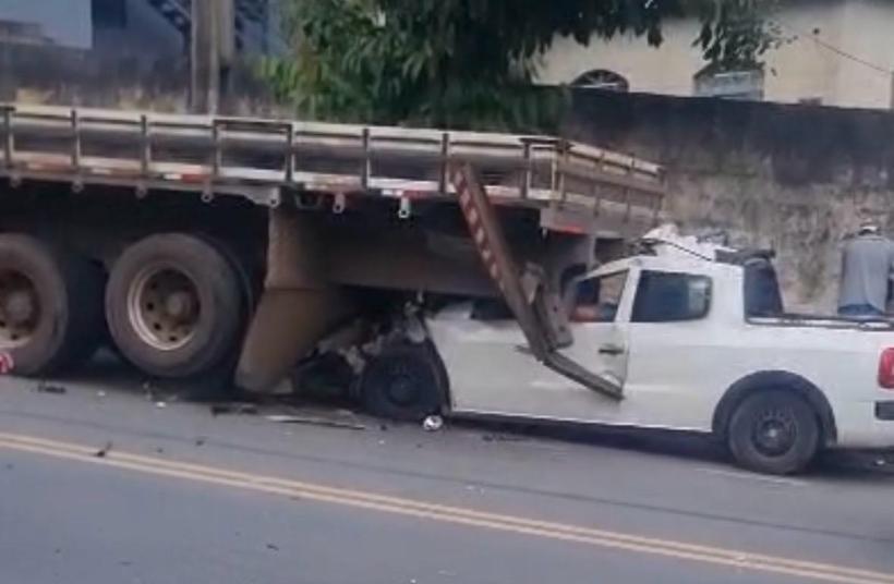 Caminhão tombado sobre um carro branco em uma rua, com danos visíveis e pessoas ao fundo avaliando a situação.