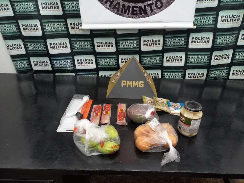 Produtos alimentícios variados dispostos sobre uma mesa, incluindo frutas, vegetais, embalagens de carne, um pote de maionese e um triângulo com a sigla PMMG ao fundo.