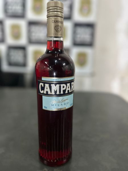 Garrafa de Campari, bebida alcoólica de cor vermelha, com rótulo em azul e branco, sobre uma superfície escura.