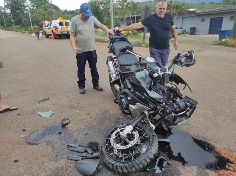 Homens observando uma motocicleta BMW danificada em uma estrada, com vazamento de óleo e destroços visíveis ao redor.