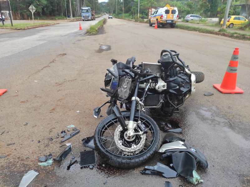 Motocicleta caída na estrada, com danos visíveis e destroços ao redor. Sinalização de segurança presente ao fundo.