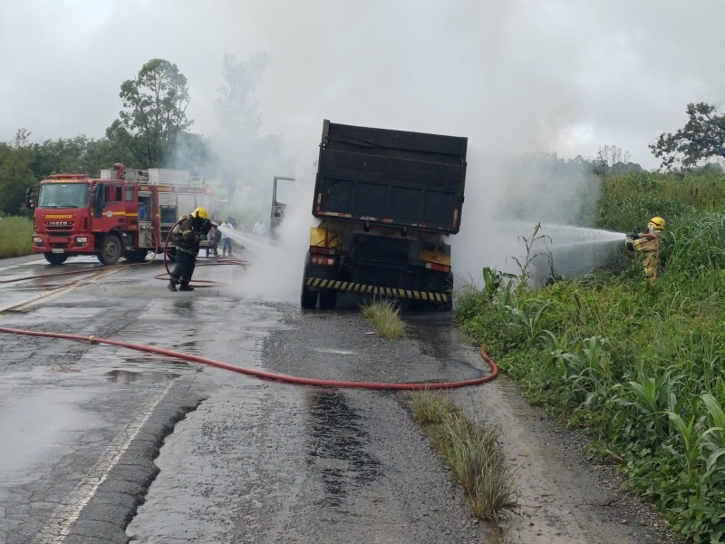 Caminhão pegando fogo na estrada, com bombeiros apagando as chamas. Há fumaça e um caminhão de bombeiros ao fundo.