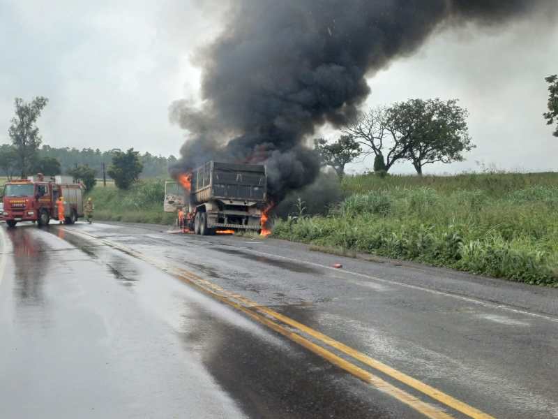 Caminhão em chamas na beira da estrada, com fumaça preta subindo e bombeiros tentando controlar o fogo.