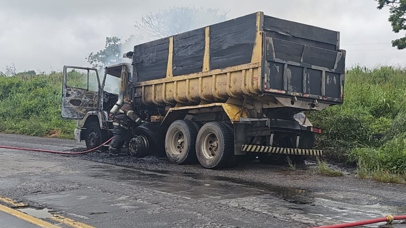 Caminhão baú pegando fogo em estrada, com bombeiros tentando apagar as chamas.