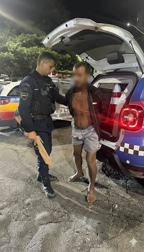 Policial interagindo com um homem descalço e sem camisa próximo a um carro de polícia à noite.