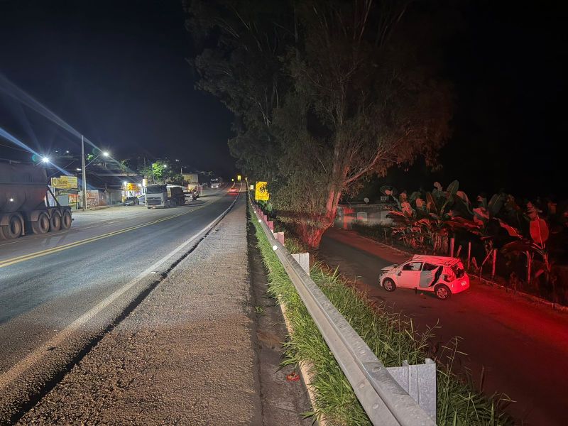 Imagem noturna de uma estrada com caminhões e um carro pequeno estacionado à direita, cercado por vegetação.