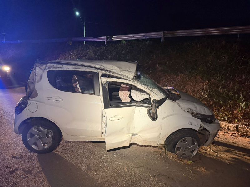 Carro branco danificado após acidente, com a porta do motorista aberta e airbags acionados, à noite.