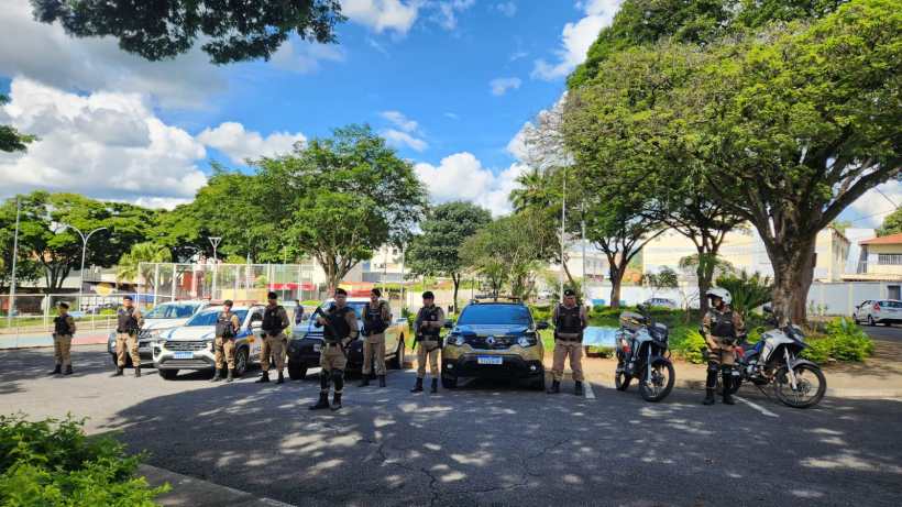 Grupo de policiais em frente a viaturas e motocicletas em um parque, com árvores ao fundo e céu azul.