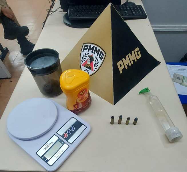 Mesa com uma balança digital, um recipiente preto, um frasco com líquido, um pote de substância, uma pirâmide decorativa com o logo da PMMG e projéteis de munição.
