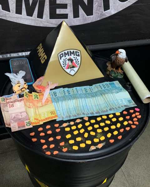 Uma mesa redonda decorada com uma pirâmide preta e dourada com o logotipo PMMG, notas de dinheiro em várias denominações, um rolo de papel, doces coloridos e uma figura de águia.