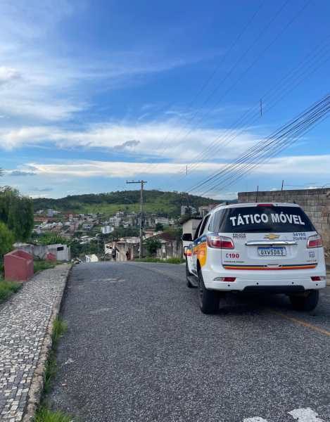 Veículo policial 'Tático Móvel' estacionado em uma estrada com vista para um bairro, rodeado por natureza e céu azul.
