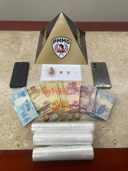 Imagem de uma mesa com objetos variados, incluindo dois celulares, notas de dinheiro, moedas, três pequenos itens sobre um papel branco e uma pirâmide de cartolina com o logo PMMG.