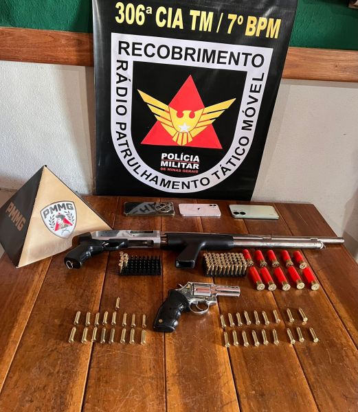 Imagem de uma mesa com armas e munições, incluindo uma espingarda, um revólver, cartuchos de munição, e um banner da Polícia Militar com o logotipo.