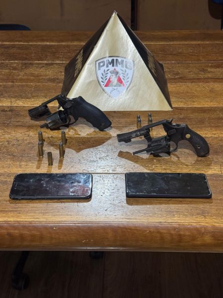 Duas pistolas sobre uma mesa de madeira, acompanhadas de cartuchos e dois celulares, com uma pirâmide de madeira ao fundo com o logo da PMMG.