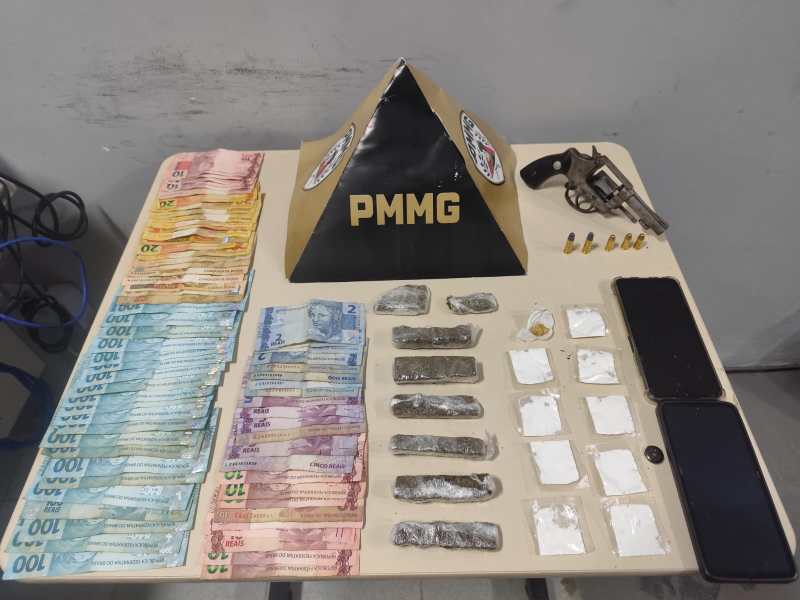 Mesa com dinheiro em notas de diferentes valores, drogas em embalagens, um revólver e dois celulares, com uma pirâmide de sinalização da PMMG ao fundo.