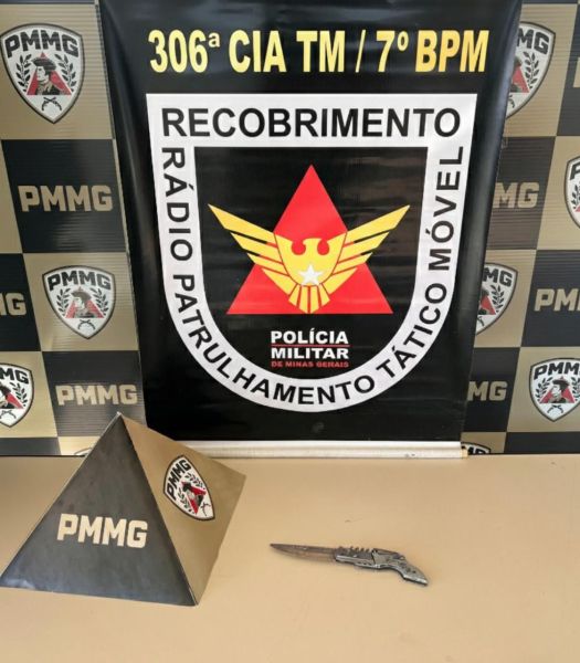 Imagem de um estande com o emblema da Polícia Militar de Minas Gerais, apresentando a sigla PMMG e a frase 'RECOBRIMENTO RÁDIO PATRULHAMENTO TÁTICO MÓVEL'. Um objeto triangular preto com a sigla PMMG está à esquerda e uma faca está posicionada em frente ao banner.