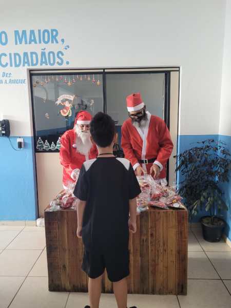 Criança de costas observando dois papais noéis em uma mesa com sacolas de presentes.