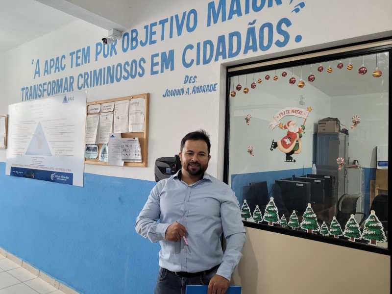 Homem em pé em um ambiente institucional com uma parede decorada com uma citação sobre transformação de criminosos em cidadãos, ao fundo uma janela decorada para o Natal.