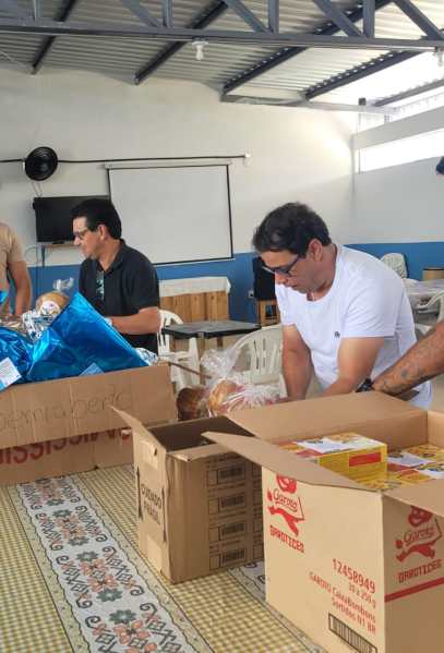 Três homens organizando alimentos em caixas em um ambiente com mesas e cadeiras.