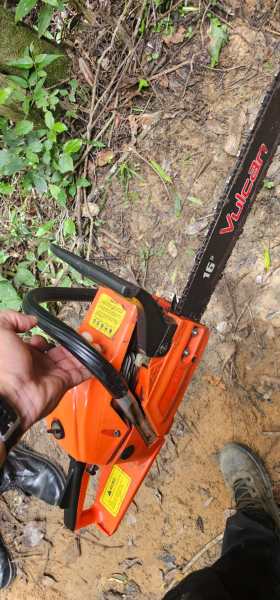 Um serrote elétrico laranja da marca Vulcan, com lâmina de 16 polegadas, segurado por uma mão, em um ambiente de floresta com folha e solo visíveis.