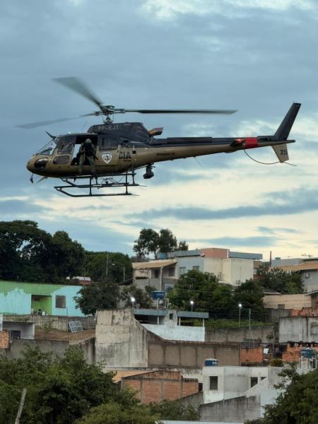 Helicóptero militar sobrevoando uma área urbana com prédios e vegetação ao fundo.