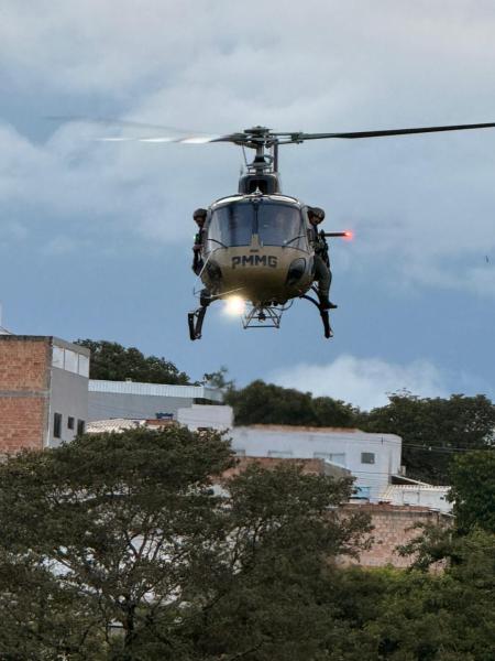 Um helicóptero da PMMG voando sobre um bairro, com dois policiais armados visíveis.